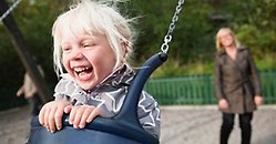 Ett gungande barn skrattar stort medan en förälder står i bakgrunden och puttar på. 