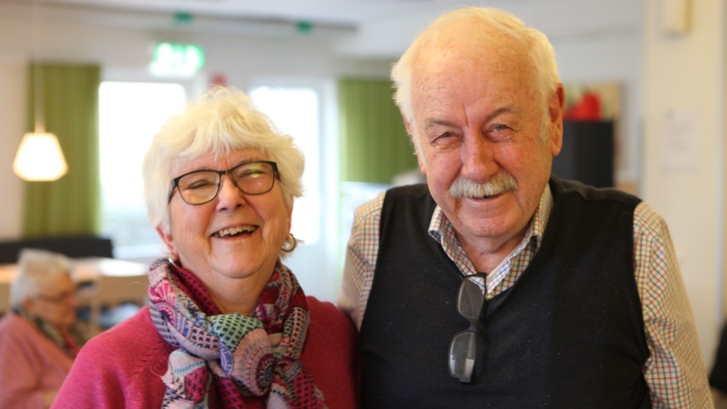 En årsrik kvinna med glasögon och vitt kortklippt hår samt en äldre herre i väst och skjorta som står bredvid varandra och skrattar på träffpunkt Norrehed i Löddeköpinge