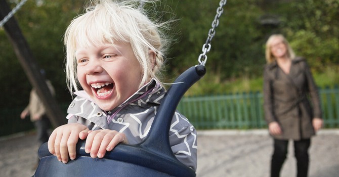 Ett gungande barn skrattar stort medan en förälder står i bakgrunden och puttar på. 