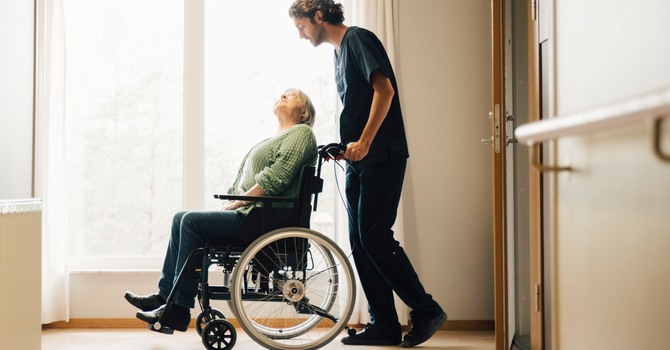 Man som arbetar på äldreboende knuffar kvinna i rullstol genom en korridor.
