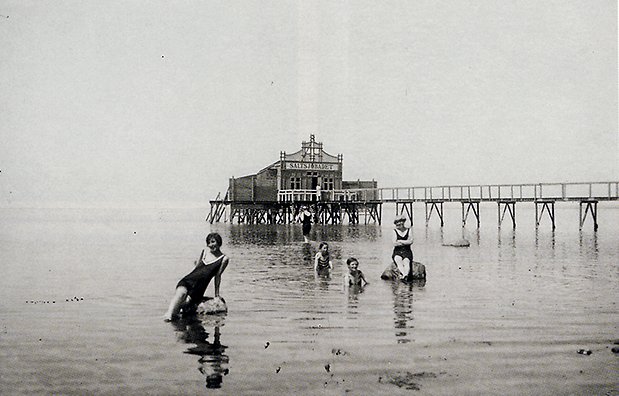 Svart vitt foto från 1910 av badande damer i havet men brygga i bakgrunden.