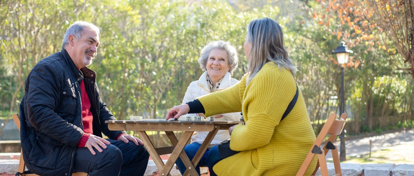 En äldre man och två äldre kvinnor sitter kring ett cafébord i trä och umgås utomhus.